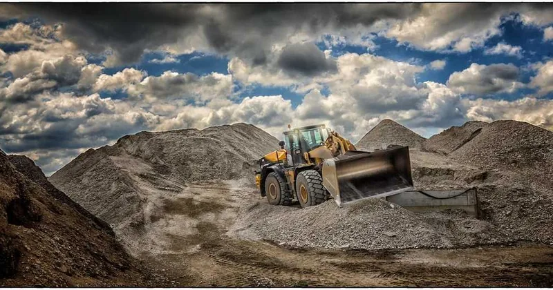 Excavateur jaune en action sur un site de construction, avec des montagnes de terre et des nuages au-dessus